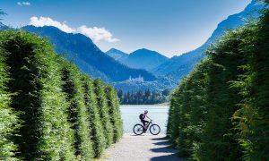 Radfahrerin auf E-Bike fährt am See entlang, dahinter Berge